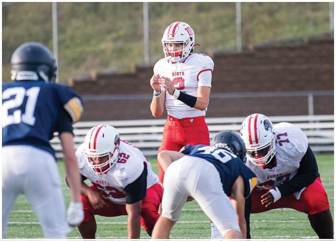 A group of people in football uniforms are positioned on a field with one person in a white and red uniform preparing to throw a football while others in blue and white uniforms are lined up opposite them. Description generated by AI
