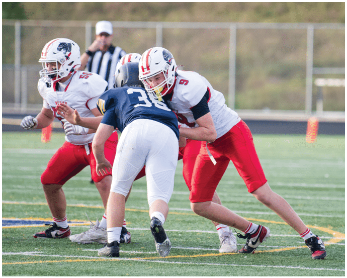 People wearing football uniforms are engaged in a play on a grassy field with a chain-link fence and blurred background. Description generated by AI