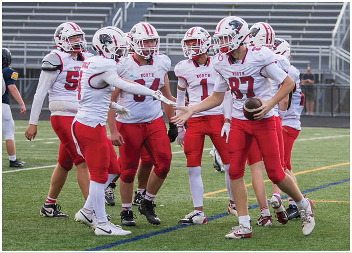 A group of people wearing white and red football uniforms are gathered on a field with one person holding a football and others appearing to celebrate or communicate. Description generated by AI