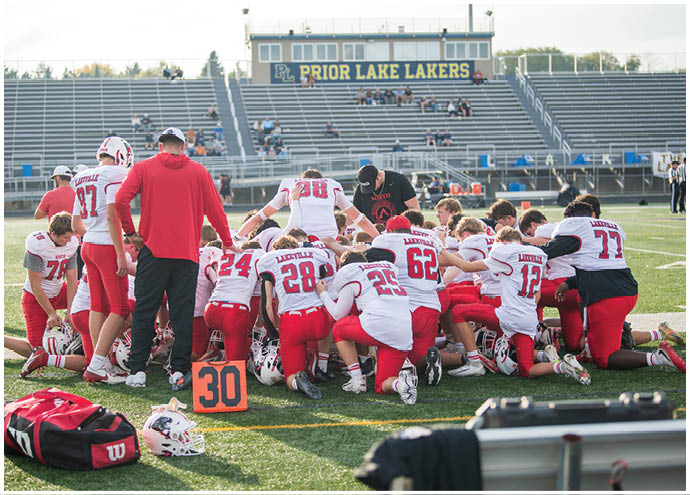 A group of football players in white and red uniforms kneel together in a huddle on a field with a coach standing nearby and a scoreboard reading “Prior Lake Lakers" in the background. Description generated by AI