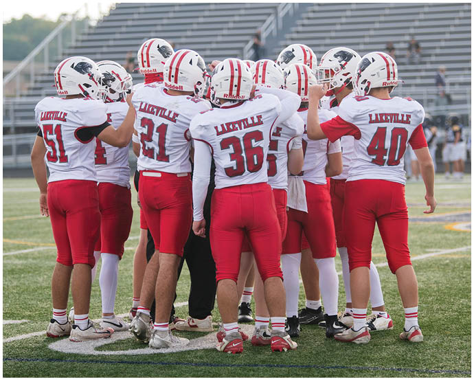 A group of people wearing red and white football uniforms with helmets are huddled together on a sports field with empty bleachers in the background. Description generated by AI