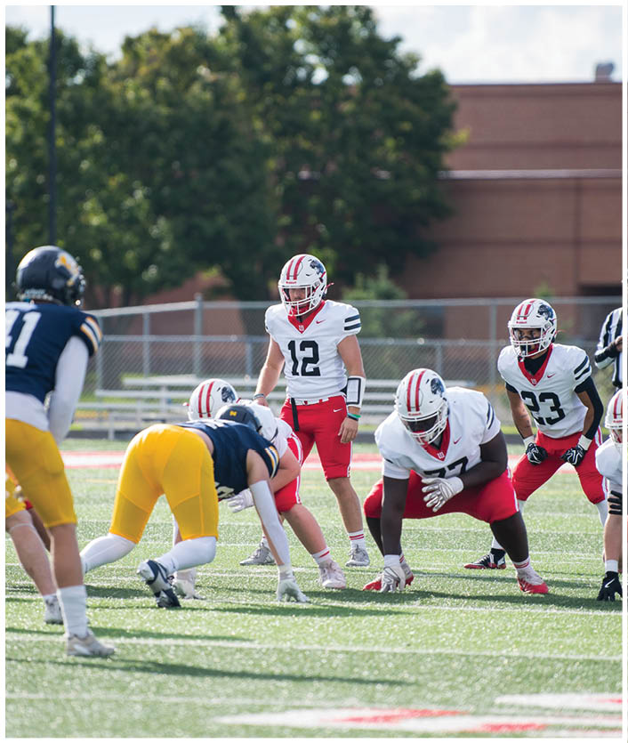 People wearing football uniforms are positioned on a field preparing for a play during a game. Description generated by AI