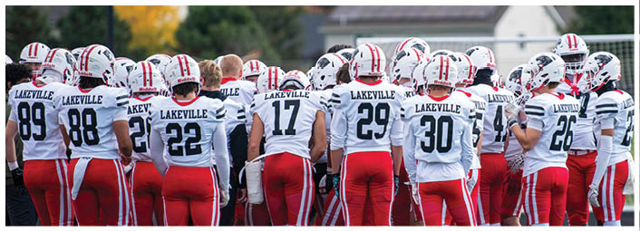 A group of people wearing white football uniforms with “Lakeville" on the back are gathered on a field near a goalpost, appearing to be in a team huddle or discussion. Description generated by AI