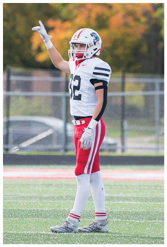 Two people in football uniforms stand on a field with one raising an arm and the other holding a hand near their face, with trees and a fence in the background. Description generated by AI