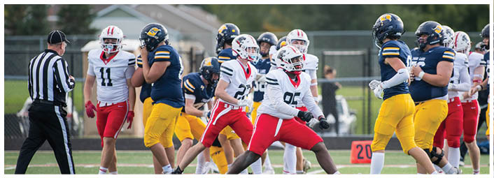 Adults in football uniforms from two teams are on a field with a referee nearby, appearing to be in the middle of a game or play. Description generated by AI