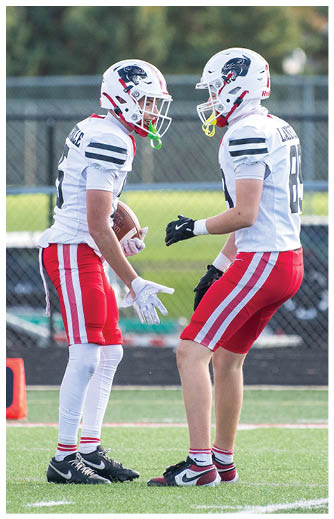 Two people wearing white football jerseys with red pants and helmets are on a football field, with one holding a football and the other reaching out with a gloved hand. Description generated by AI