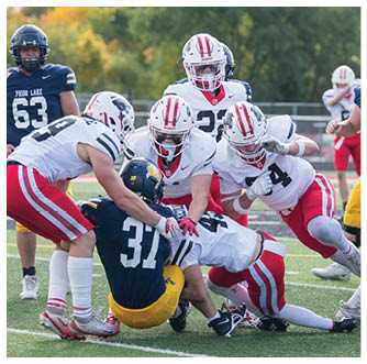 A group of people wearing football uniforms are actively engaged in a play on a grassy field with some players in white and red uniforms tackling or blocking players in navy blue and yellow uniforms. Description generated by AI