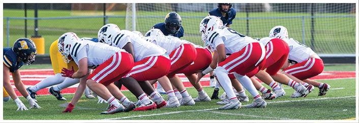 Two opposing American football teams are lined up at the line of scrimmage on a green field with one team in white and red uniforms and the other in navy blue and yellow uniforms. Description generated by AI