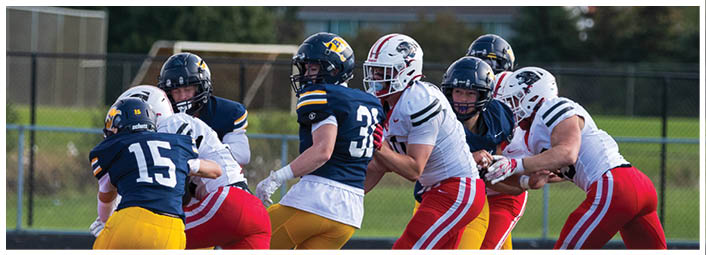 People wearing football uniforms are engaged in a play on a grassy field with trees and houses in the background. Description generated by AI