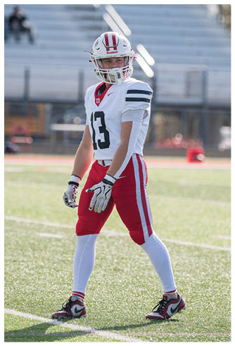 A person in a white football uniform with red pants and a helmet stands on a sunlit football field with empty bleachers and a referee in the background. Description generated by AI