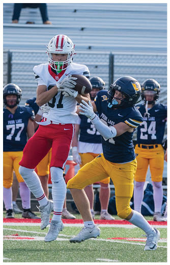 A person wearing a white football uniform with red pants and the name Lakeville on the back is running on a football field while three people in black and yellow uniforms stand in the background. Description generated by AI