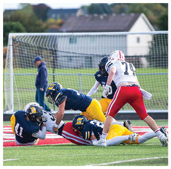 Several people in football uniforms are engaged in a play on a field near a goalpost with a person standing in the background. Description generated by AI