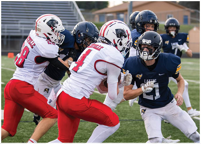 People in football uniforms are actively engaged in a play on a grassy field with one player in a dark jersey attempting to evade players in white and red uniforms. Description generated by AI