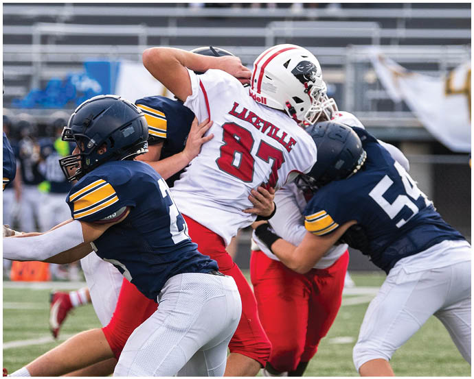 People in football uniforms are engaged in a tackle during a game on a field with empty bleachers in the background. Description generated by AI