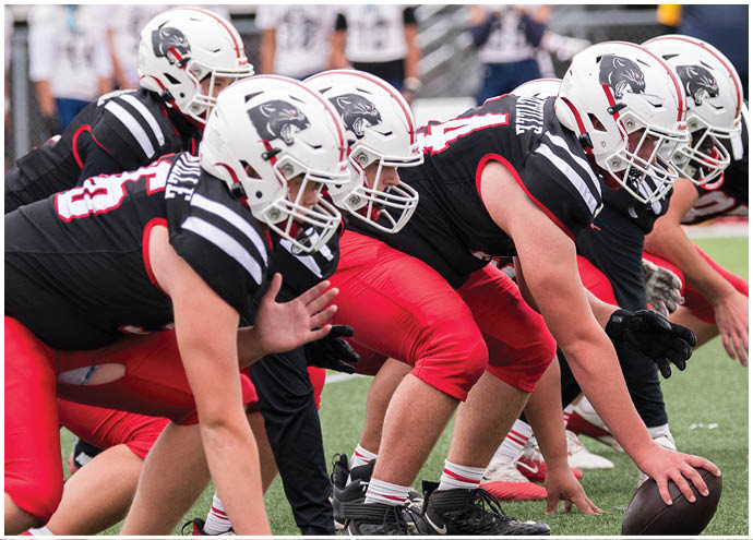 A group of people in football uniforms and helmets are lined up on a field in a crouched position, preparing for a play. Description generated by AI