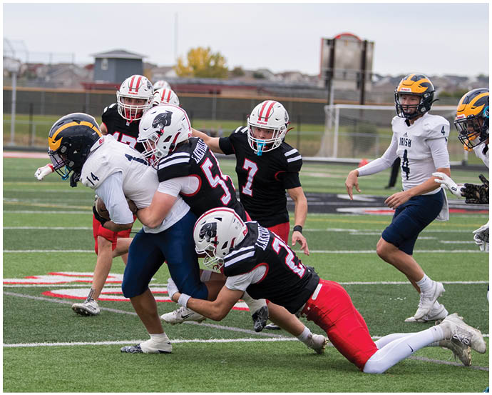 Players in football uniforms are actively engaged in a play on a field with one player in a white jersey preparing to move while others in black and red jerseys are positioned around him. Description generated by AI