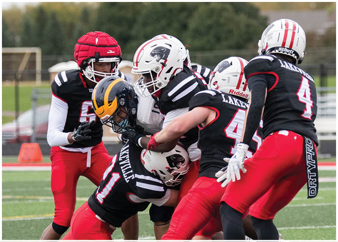 Five people in football uniforms are engaged in a tackle on a green field with a blurred background of trees and houses. Description generated by AI