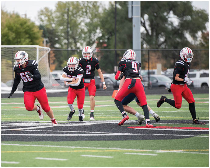 Several people wearing red pants and black jerseys are playing football on a field with a goalpost and cars visible in the background. Description generated by AI