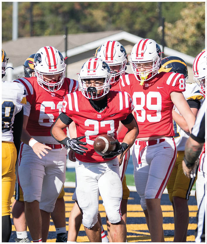 A group of football players in red and white uniforms and others in white and yellow uniforms are gathered on a field near the end zone with a referee standing nearby. Description generated by AI