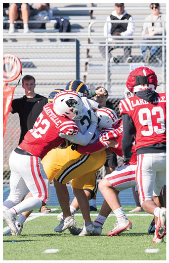 Three football players in red and white uniforms tackle a player in a yellow and blue uniform on a field with bleachers in the background. Description generated by AI