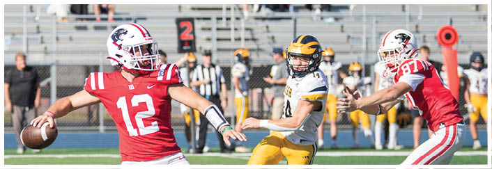 A person in a red football uniform with the number 12 is preparing to throw a football while another person in a yellow and black uniform is running toward them on a football field. Description generated by AI