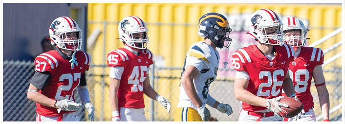 Four people wearing football uniforms are standing on a field, with one holding a football and another wearing a different colored uniform. Description generated by AI