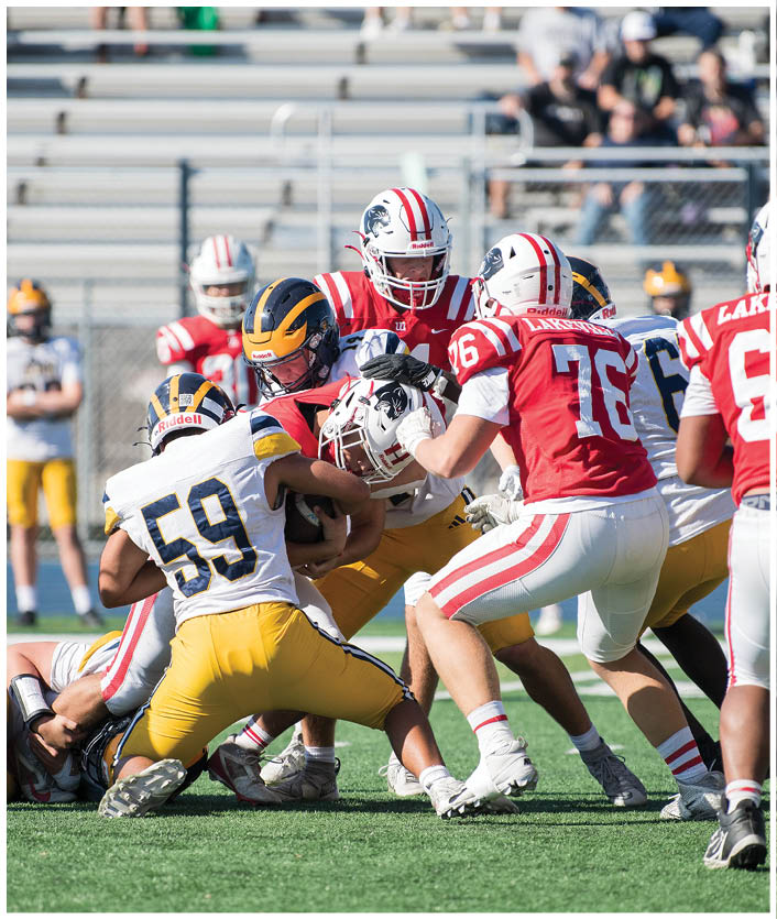 People in red and white football uniforms are engaged in a play against people in yellow and black uniforms on a green field with bleachers and spectators in the background. Description generated by AI
