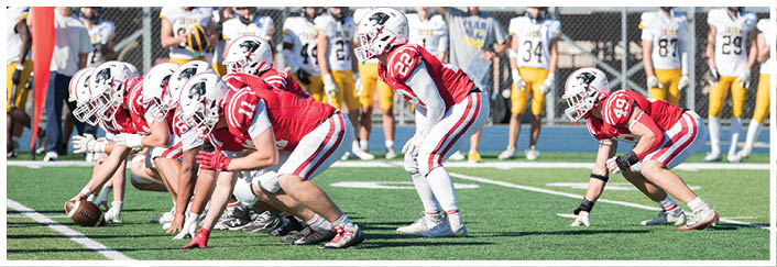 Two American football teams are lined up facing each other on a field with one team in red uniforms preparing to snap the ball and the other team in white and yellow uniforms standing in the background. Description generated by AI