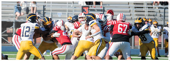People in football uniforms are engaged in a play on a field with bleachers and spectators in the background. Description generated by AI