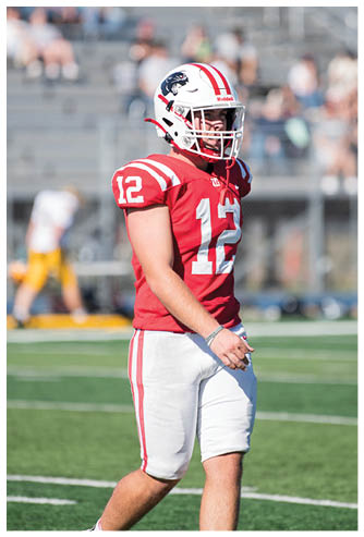 A person wearing a red and white football uniform with the number 12 stands on a football field with blurred spectators in the background. Description generated by AI