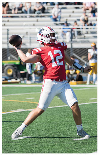 A person in a red sports jersey and white pants is on a football field with three people in yellow pants and white jerseys standing near the bleachers in the background. Description generated by AI