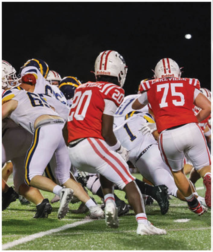 Several football players in red and white uniforms are actively engaged in a play on a well-lit field during a nighttime game. Description generated by AI