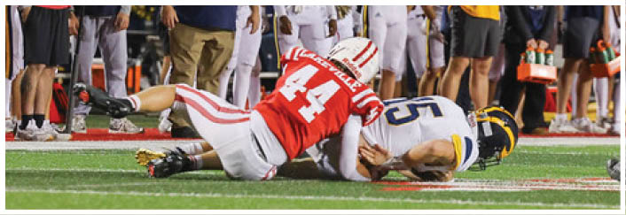 A person in a red and white football uniform is kneeling on the field while several people in white football uniforms and casual clothing stand nearby. Description generated by AI