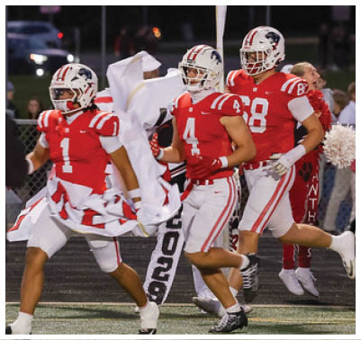 People wearing sports uniforms and cleats are running on a turf field near a red boundary line. Description generated by AI