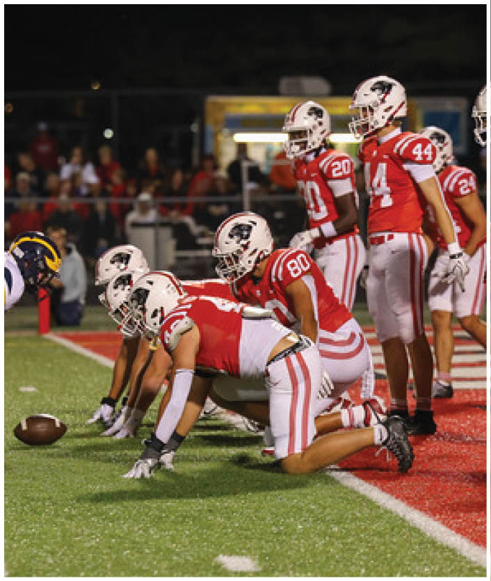 Players in red and white uniforms and players in blue and yellow uniforms are lined up facing each other on a football field, preparing for a play. Description generated by AI