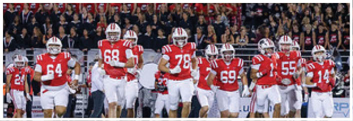A group of people wearing red and white football uniforms are running onto a field with a crowd of spectators in the background. Description generated by AI