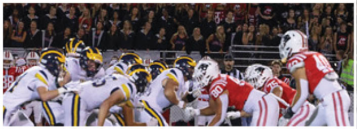 Two opposing American football teams in helmets and uniforms are lined up facing each other on the field during a game with a crowd in the background. Description generated by AI