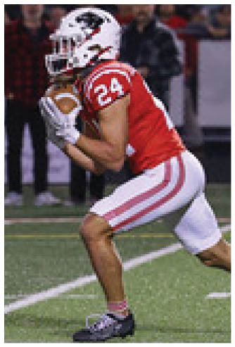 A person in a red and white football uniform is holding a football and appears to be preparing to run on a turf field with a blurred crowd in the background. Description generated by AI