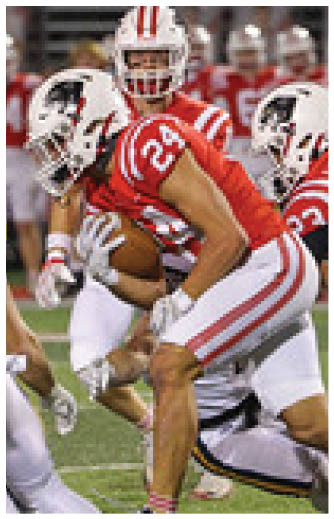 A person wearing a red and white football uniform is holding a football while being tackled by another player on a football field. Description generated by AI