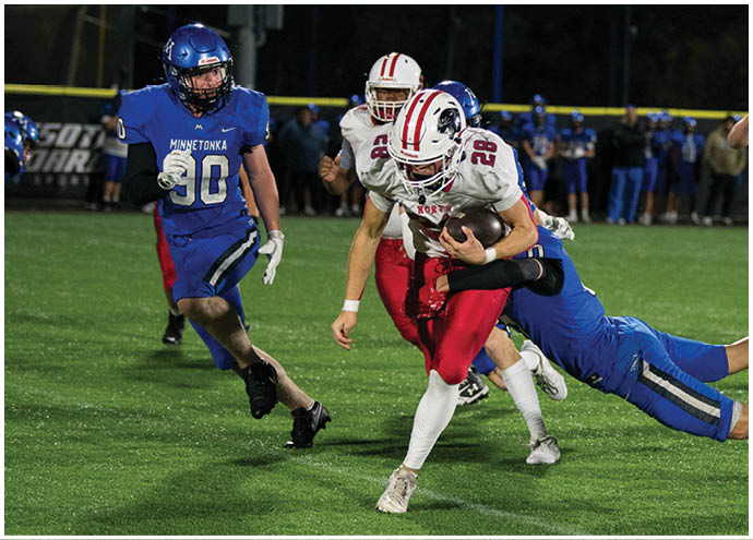 A person in a white and red football uniform is running with the ball while being tackled by a person in a blue uniform on a grassy field during a game. Description generated by AI