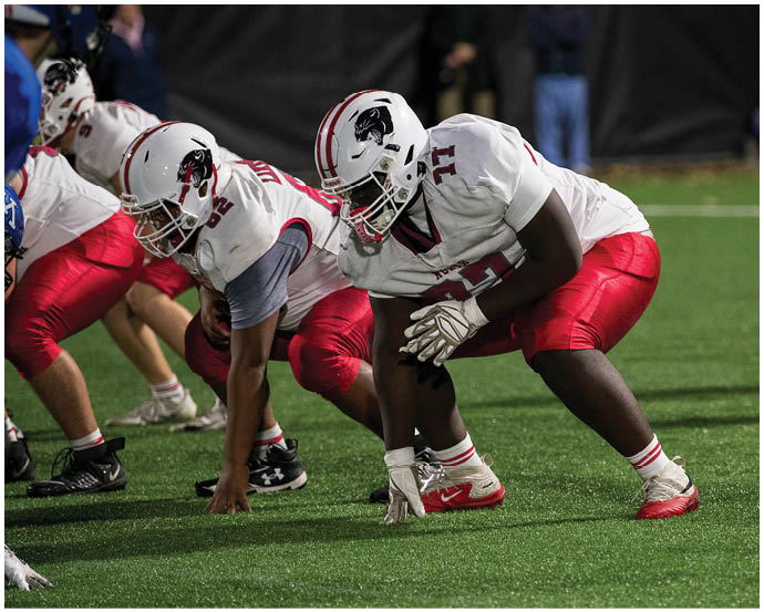 Several football players in white helmets and red pants are crouched in a ready position on a green field, preparing for a play. Description generated by AI