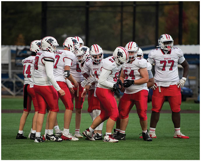 A group of people wearing white helmets, white jerseys, and red pants are gathered on a sports field, appearing to be in a team huddle or discussion. Description generated by AI