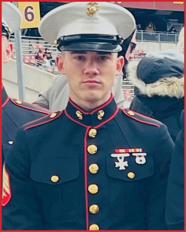 A person in a formal military dress uniform with medals stands in an outdoor setting with other people and stadium seating in the background. Description generated by AI