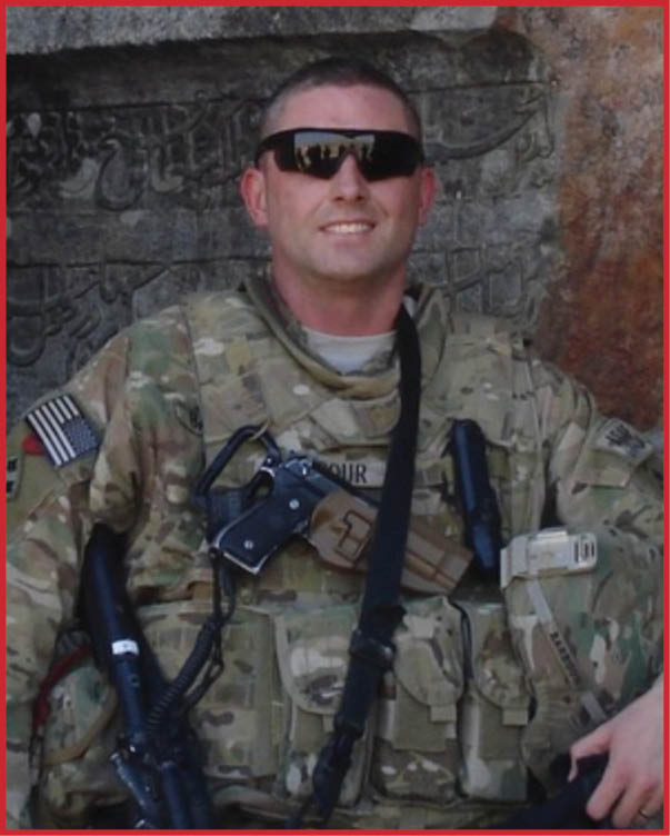 Person wearing military camouflage uniform and sunglasses holding a rifle and a helmet while standing in front of a stone wall with carved inscriptions. Description generated by AI