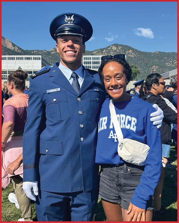 A person in a formal military uniform stands smiling with an arm around another person wearing a blue Air Force sweatshirt and shorts at an outdoor event with mountains and a crowd in the background. Description generated by AI