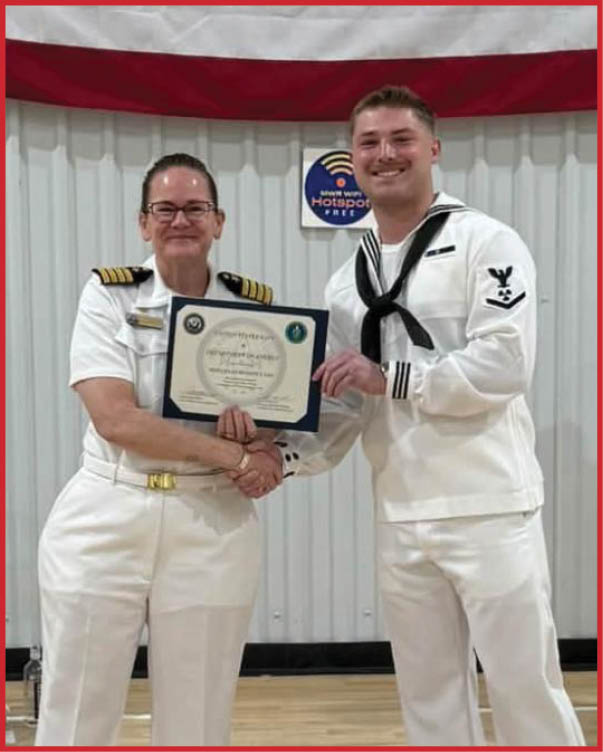 Two people in white naval uniforms stand on a gym floor under a large American flag, with one person holding a certificate while another person in uniform stands at a podium in the background. Description generated by AI