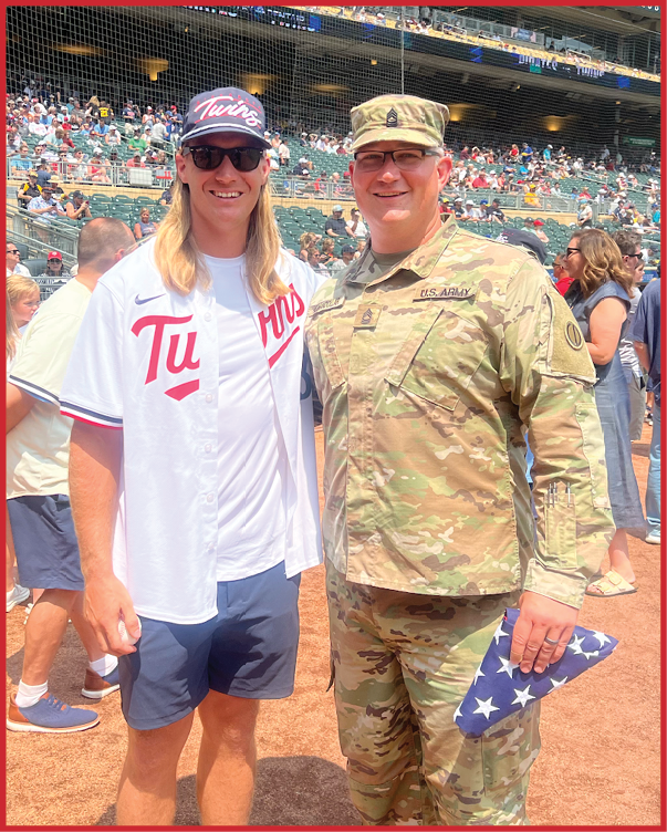 Two adults stand on a baseball field near the dugout with one wearing a military uniform and holding a folded American flag while the other wears a baseball jersey and cap. Description generated by AI