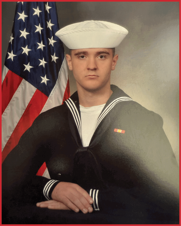 Person wearing a traditional navy sailor uniform and white hat posed with folded hands in front of an American flag. Description generated by AI