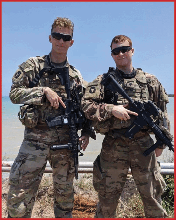 Two adults wearing camouflage military uniforms and sunglasses stand side by side holding rifles near a body of water under a clear blue sky. Description generated by AI