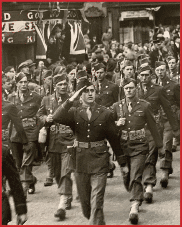 A large group of people in military uniforms is marching in formation down a street while a crowd watches and a band plays music. Description generated by AI